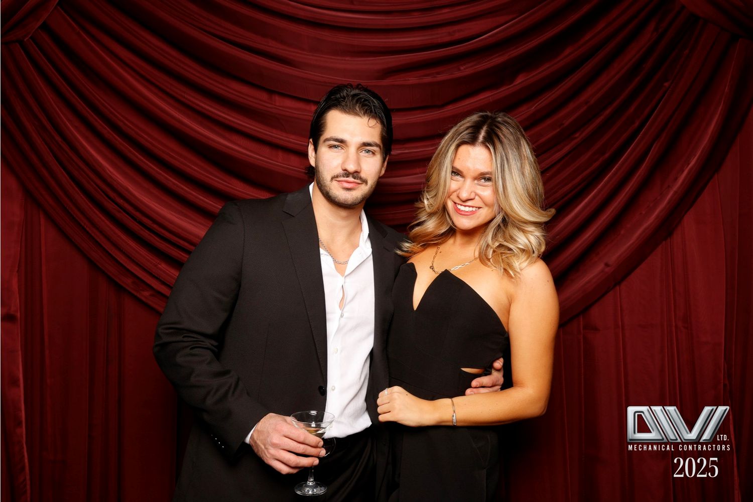 A couple in elegant evening attire poses against a red velvet curtain background.