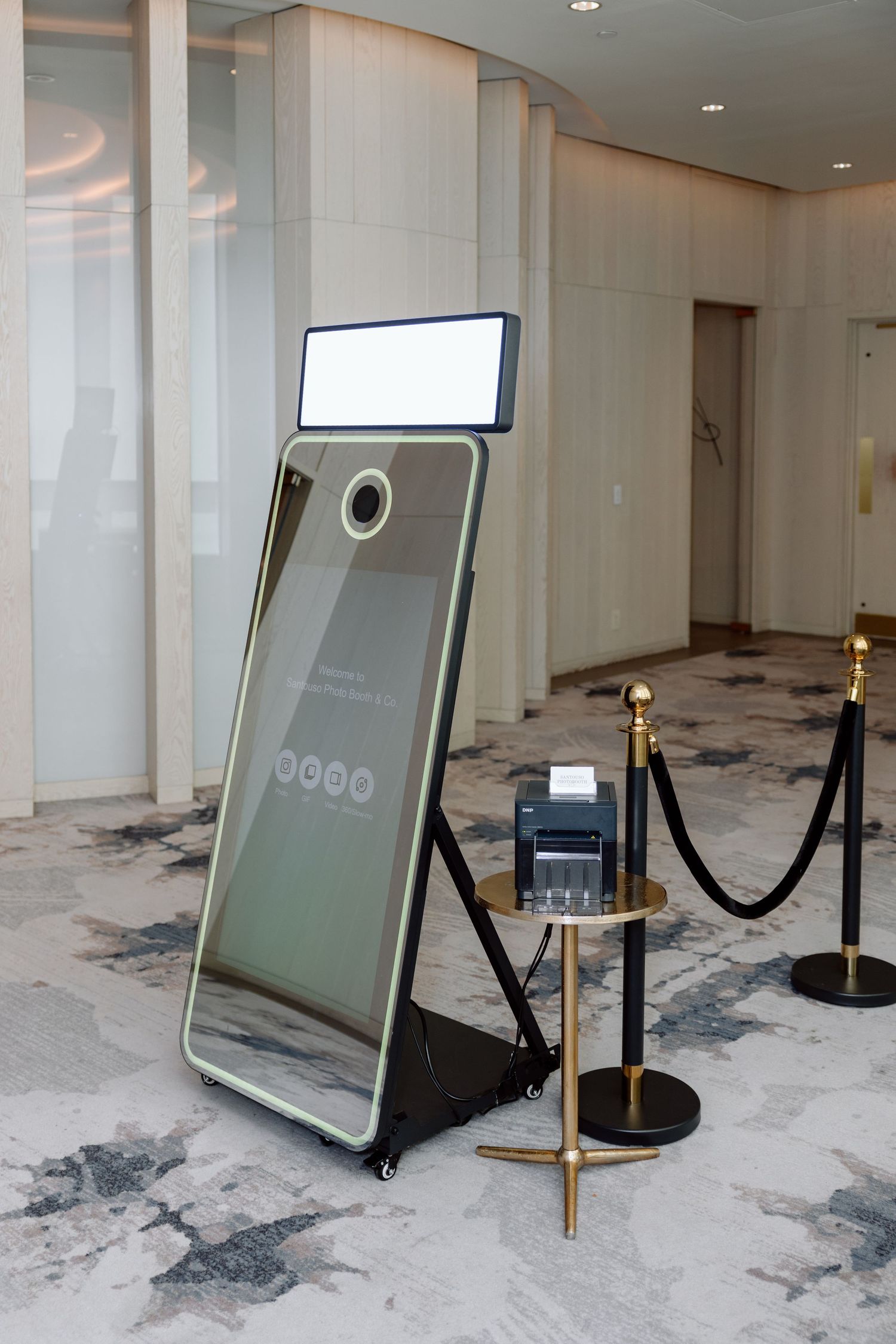 Modern mirror photo booth with LED light panel and gold accents, set up in an elegant hotel lobby with velvet rope barriers.