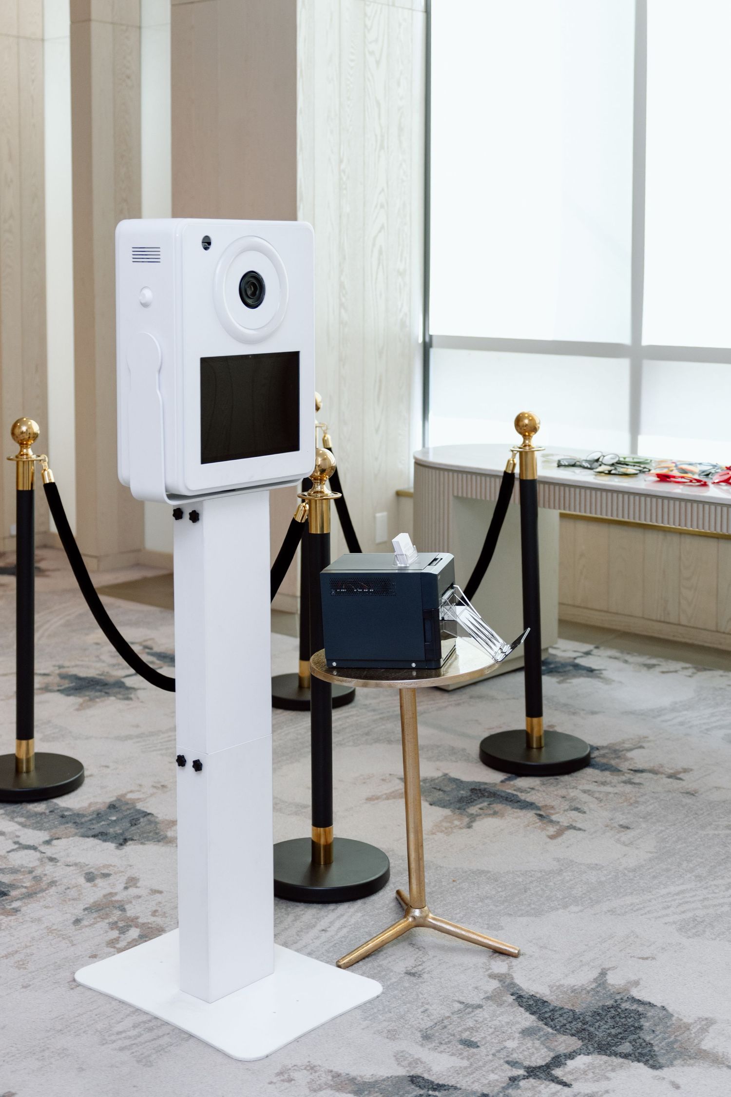 Modern white photo booth on stand with screen, surrounded by gold stanchions and velvet ropes in elegant venue.