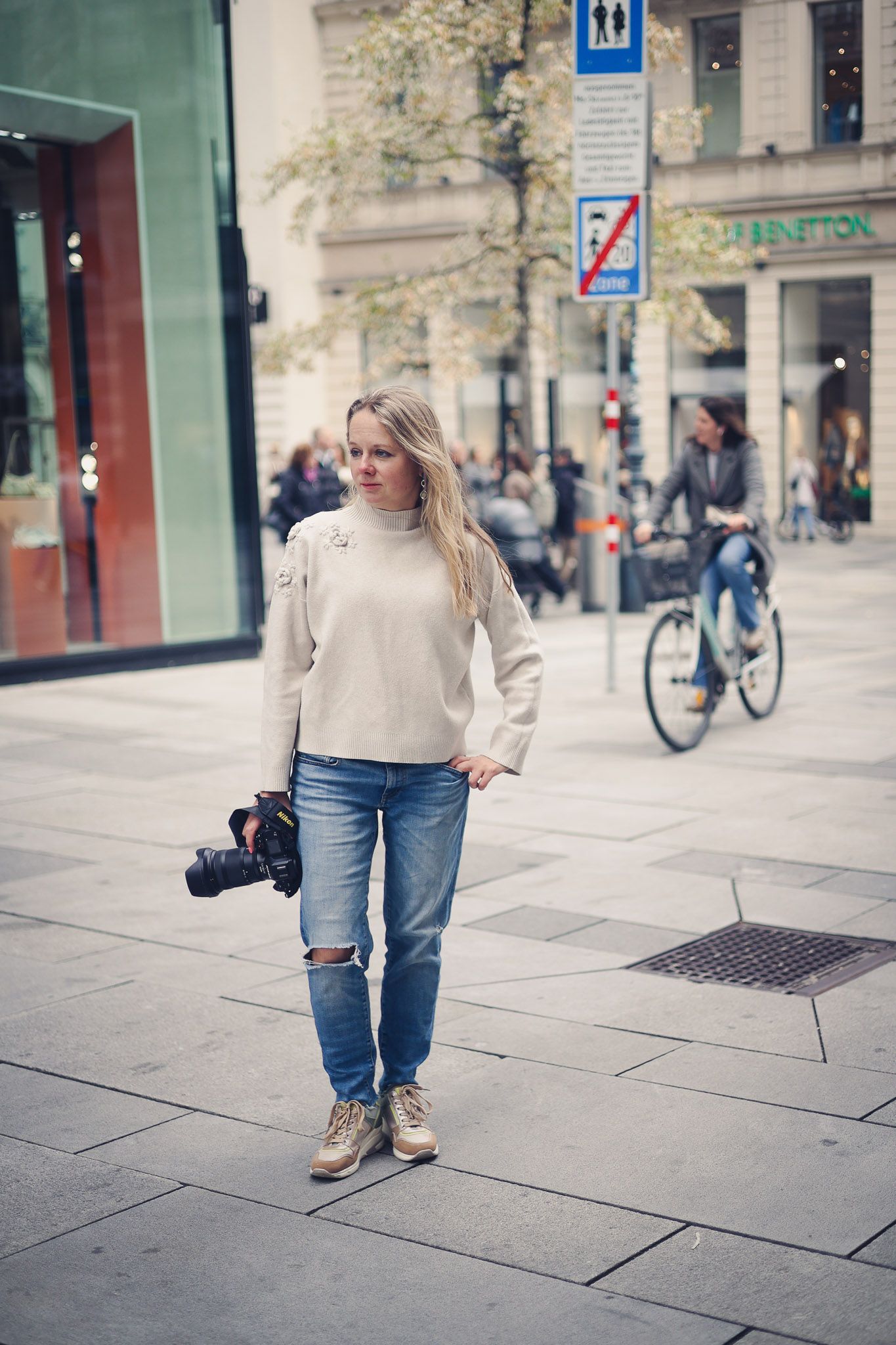 Young blonde woman holding a camera in ripped jeans and cream sweater standing on a European city street.