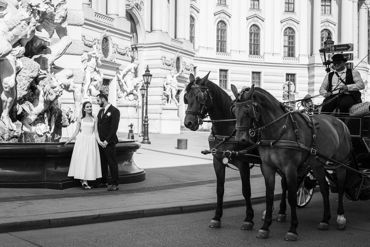 Black and white photo of a wedding couple posing near a fountain with a horse-drawn carriage in a grand European square.
