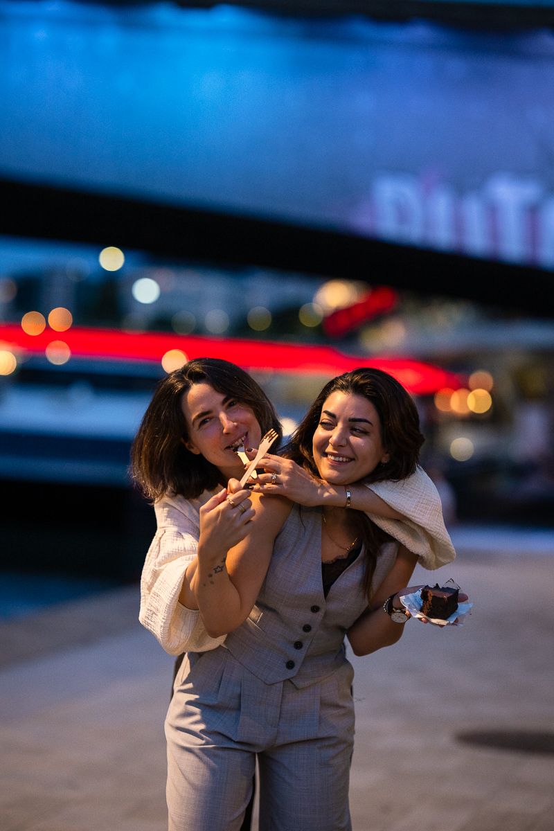 Two young women hugging and smiling outdoors at night with colorful city lights in the background.