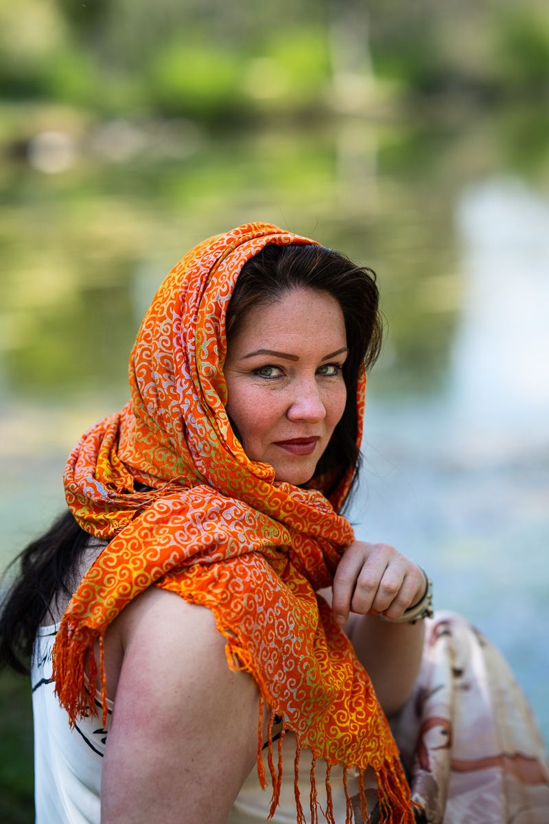 Woman in orange patterned headscarf poses outdoors near water with green foliage background.