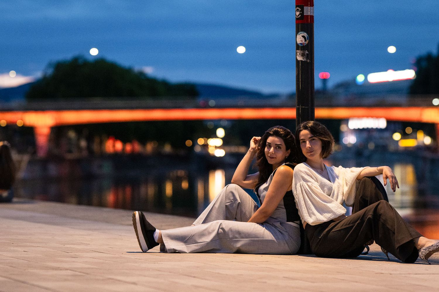 Two young women sit back-to-back on a waterfront promenade at dusk, with a glowing bridge reflected on the river.