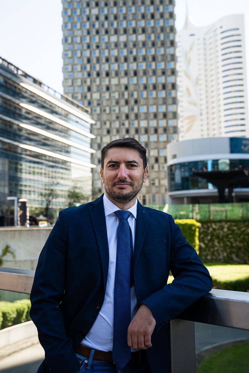 Professional businessman in navy suit and blue tie posing outdoors with modern glass skyscrapers in background.
