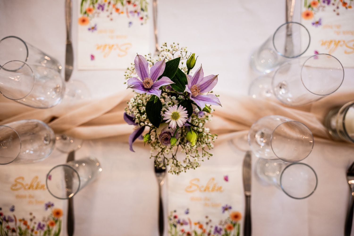 Elegant wedding table setting with purple lily and daisy bouquet, crystal glasses, and floral menu cards.