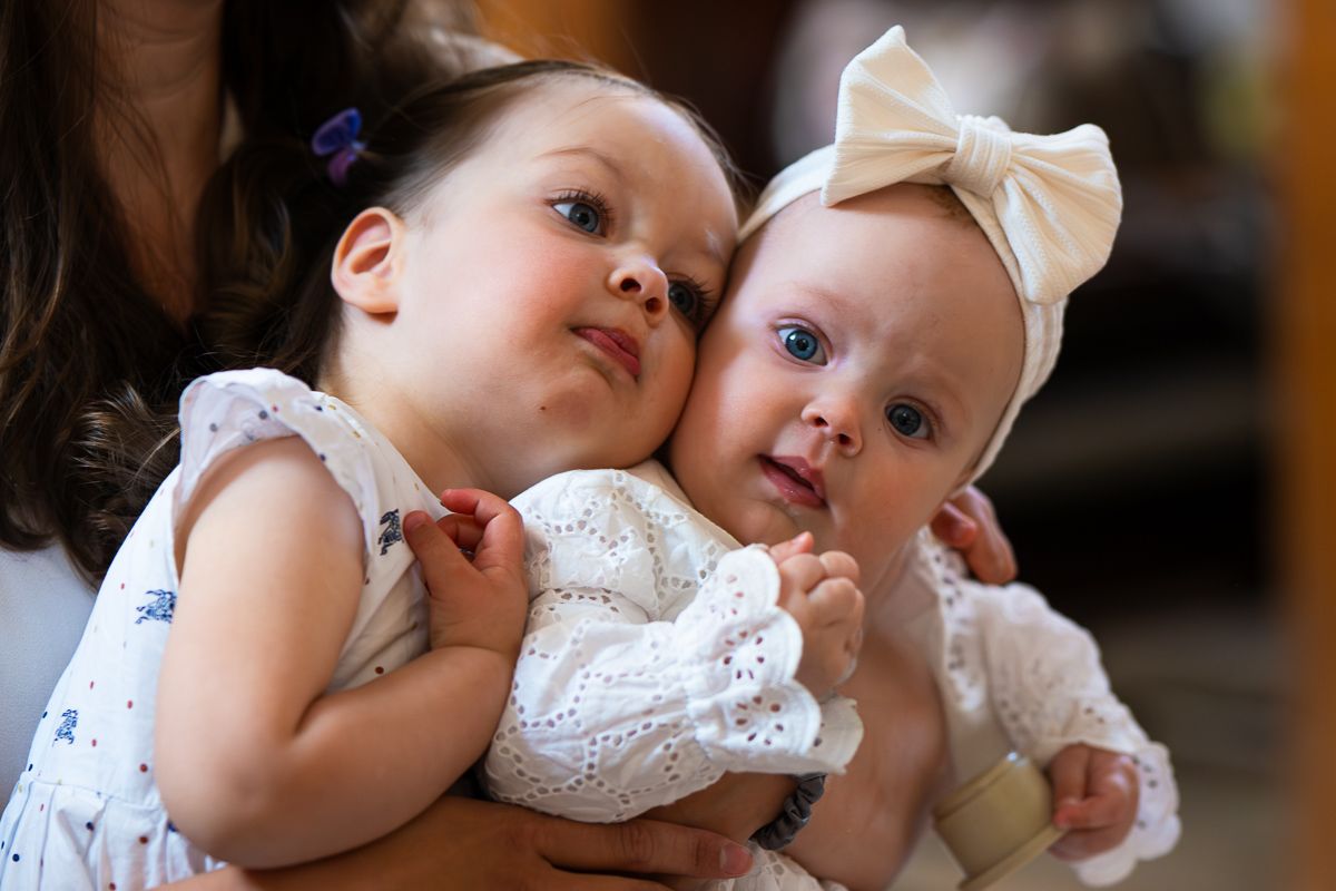Two adorable baby girls in white dresses, one hugging the other wearing a bow headband, sharing a sweet moment.