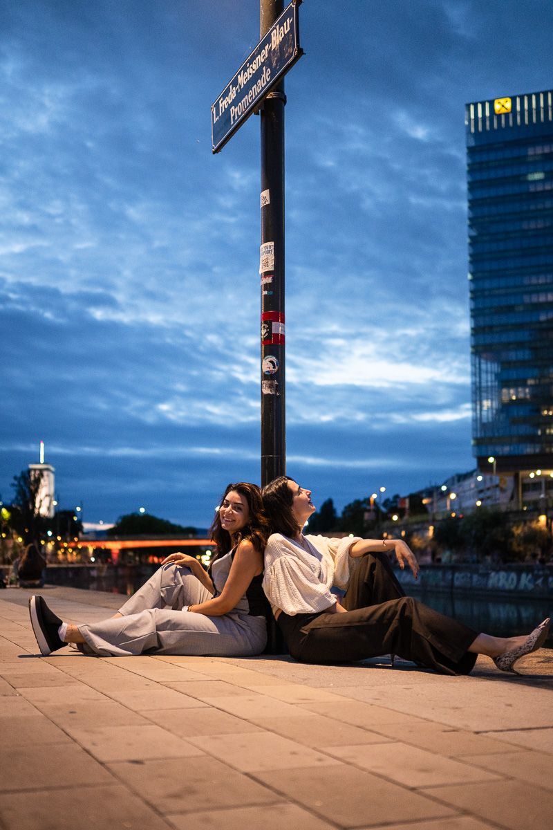 Two young women sitting on city pavement at dusk, leaning against a street sign post with urban skyline behind them.
