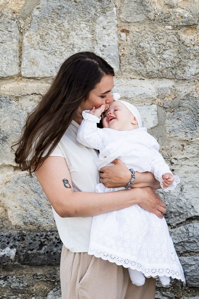A mother with long dark hair tenderly holds and nuzzles her laughing baby dressed in a white christening gown.