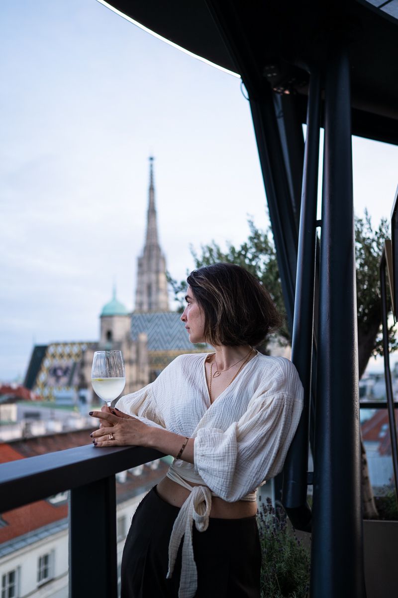 Woman holding white wine glass on rooftop balcony with St. Stephens Cathedral spire visible in Vienna skyline.
