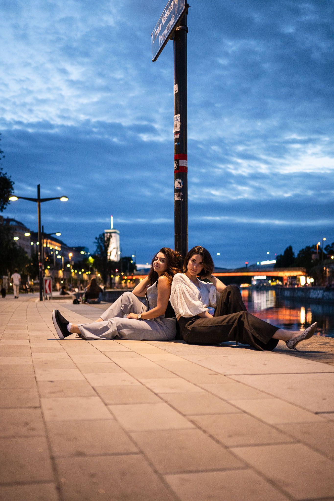 Two young women sitting back-to-back on a city sidewalk at dusk, with glowing street lights and a blue cloudy sky.