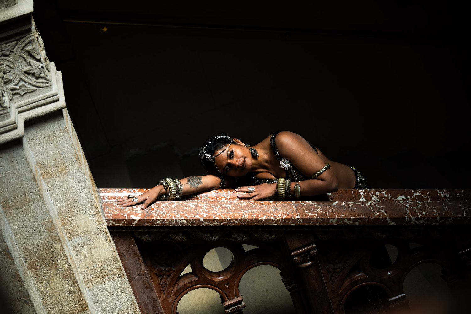 A woman in jewelry leans gracefully over a marble surface in dramatic lighting.