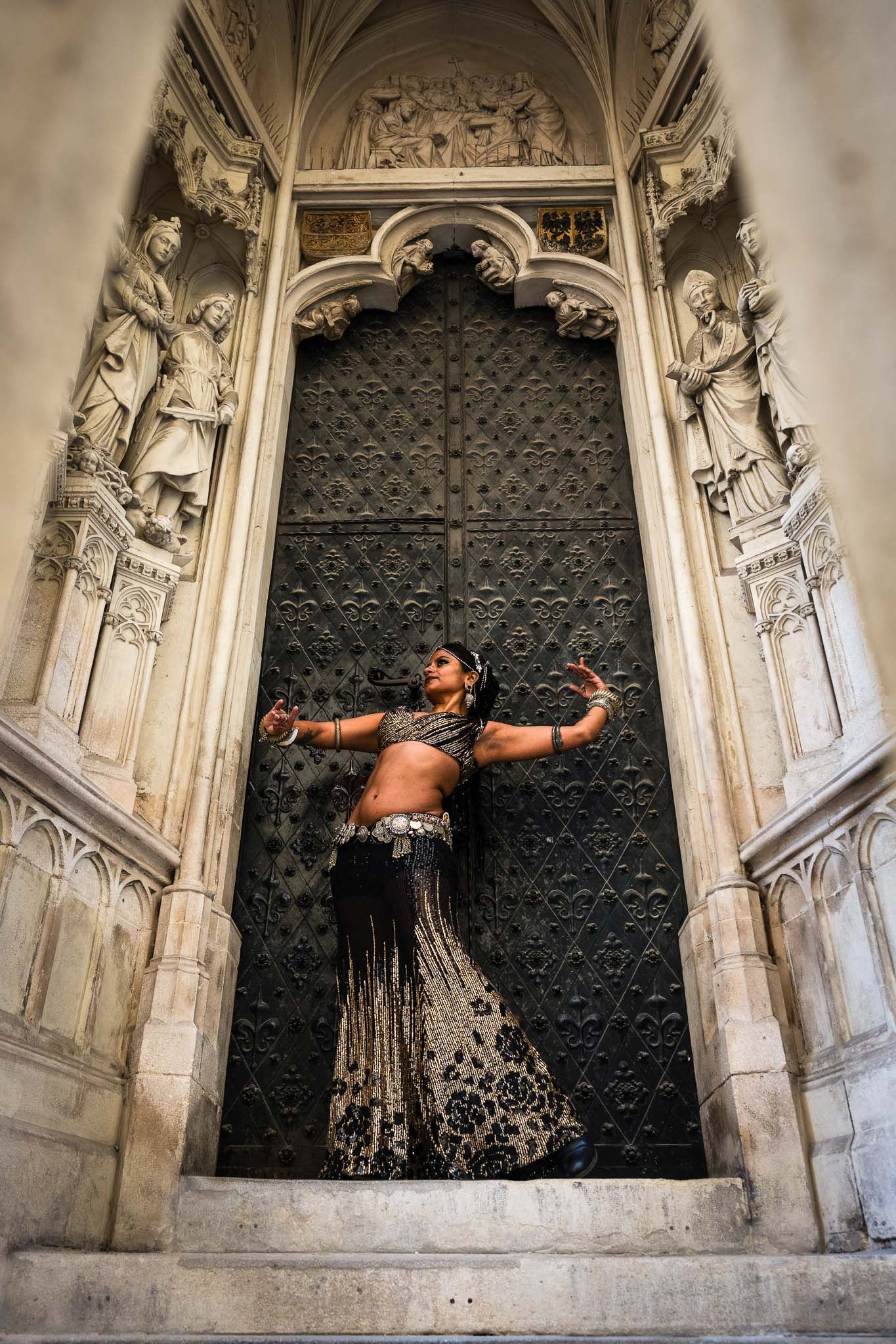 Belly dancer performing in ornate costume before grand Gothic cathedral doors with stone sculptures.