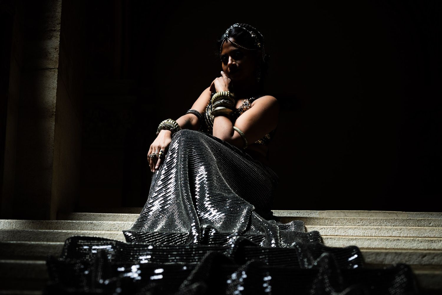Woman in silver sequin skirt and dark accessories sits on steps surrounded by black objects in dramatic lighting.