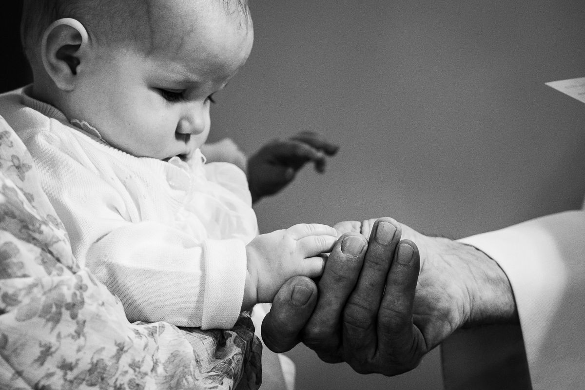 Black and white photo of a baby in white christening gown holding an elderly persons hand during baptism.