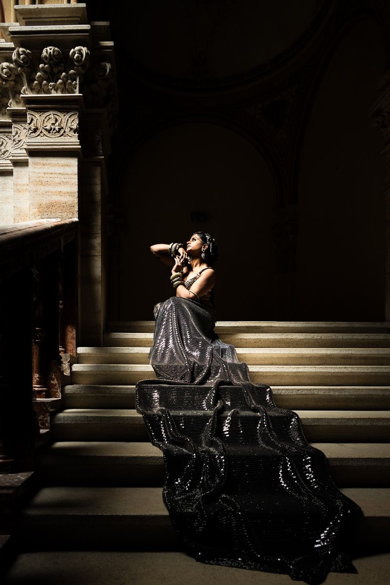 Woman in black sequin gown seated on grand staircase, gazing upward in dramatic lighting.