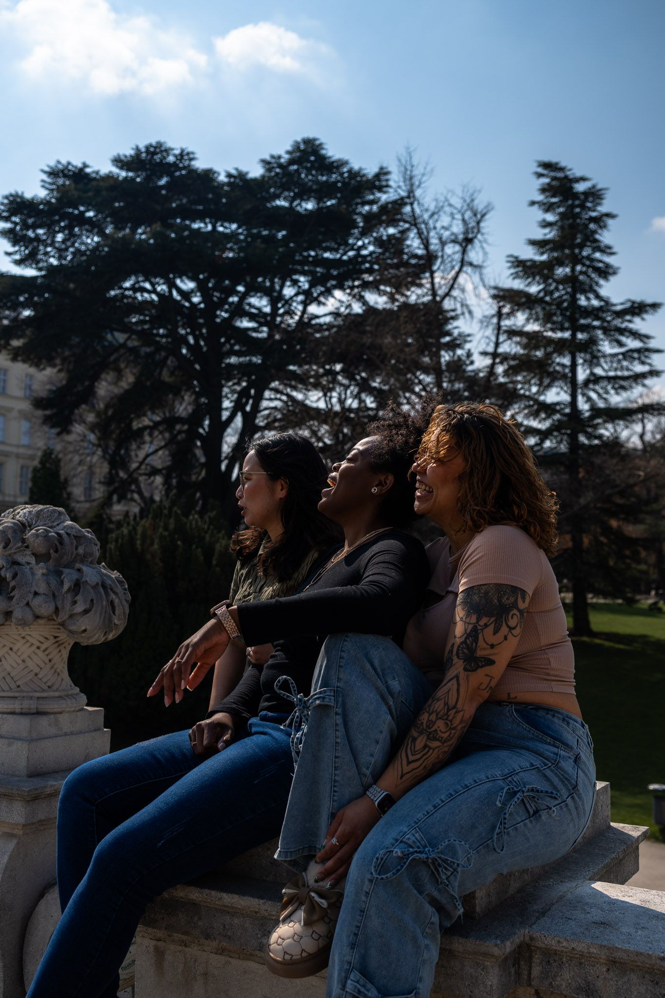 Three young women sit on a stone ledge in a sunlit park, sharing a joyful moment.