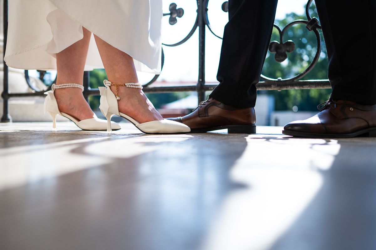 Close-up of bride's white heels and groom's brown dress shoes on a sunlit floor at a wedding.