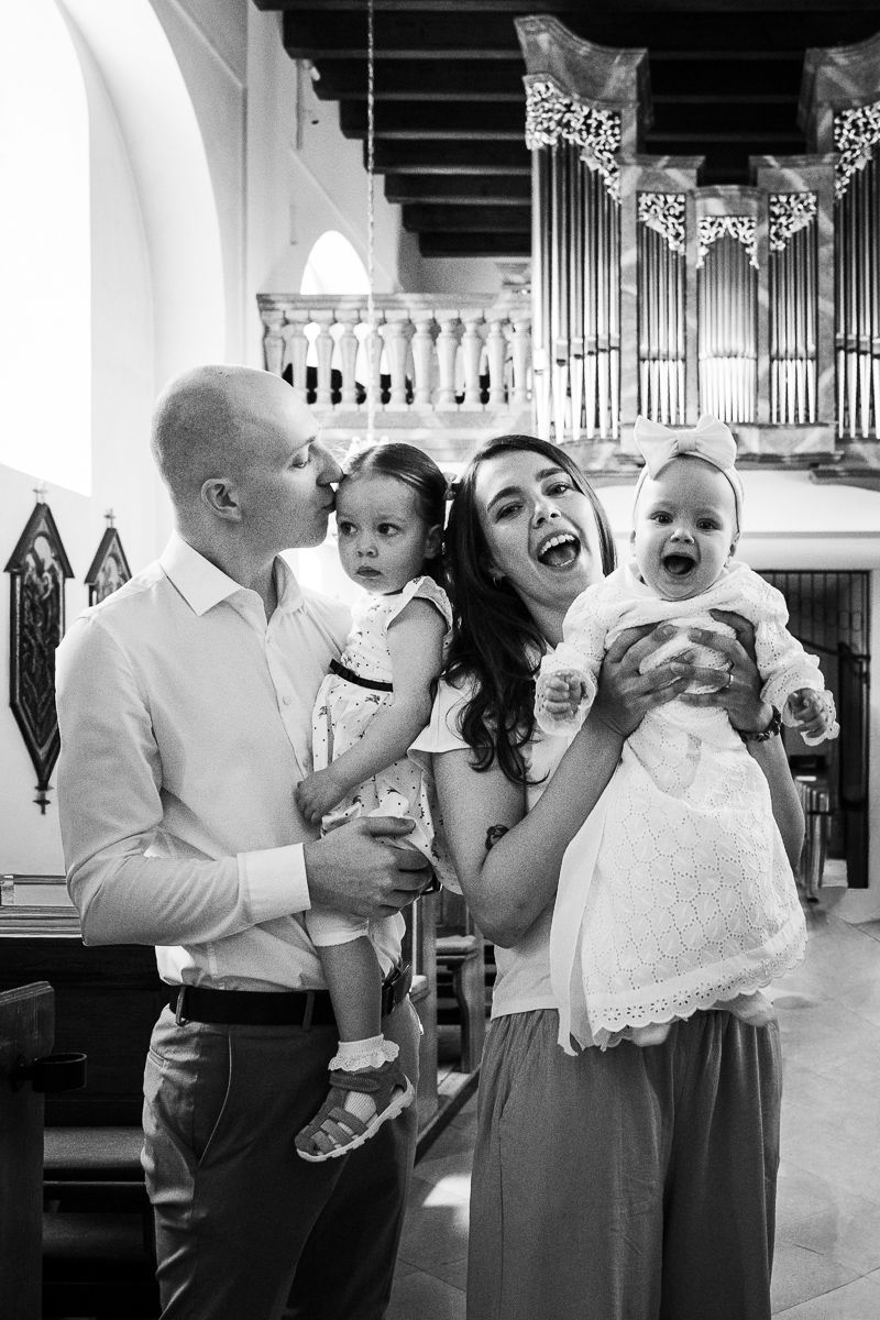 Black and white photo of a joyful family at a church baptism, parents holding two young children, smiling and laughing.