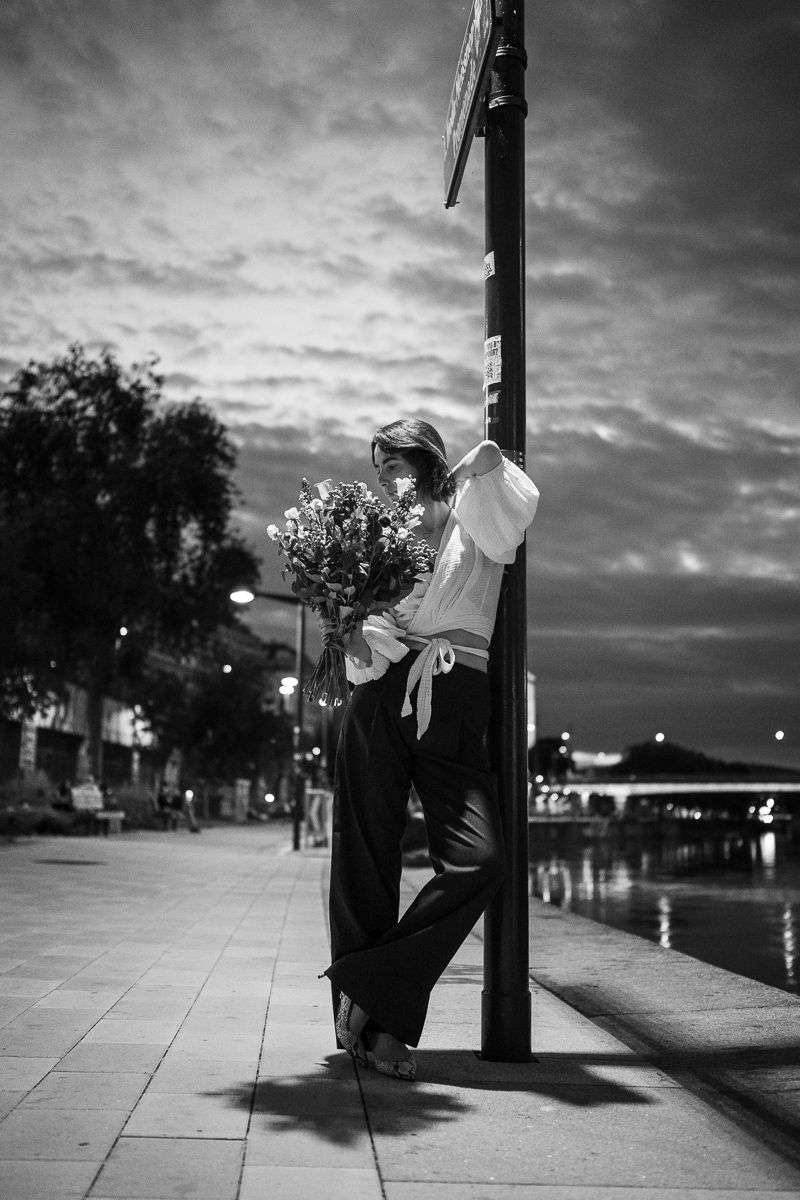 Black and white photo of a woman leaning on a street pole holding flowers at dusk on a city sidewalk.