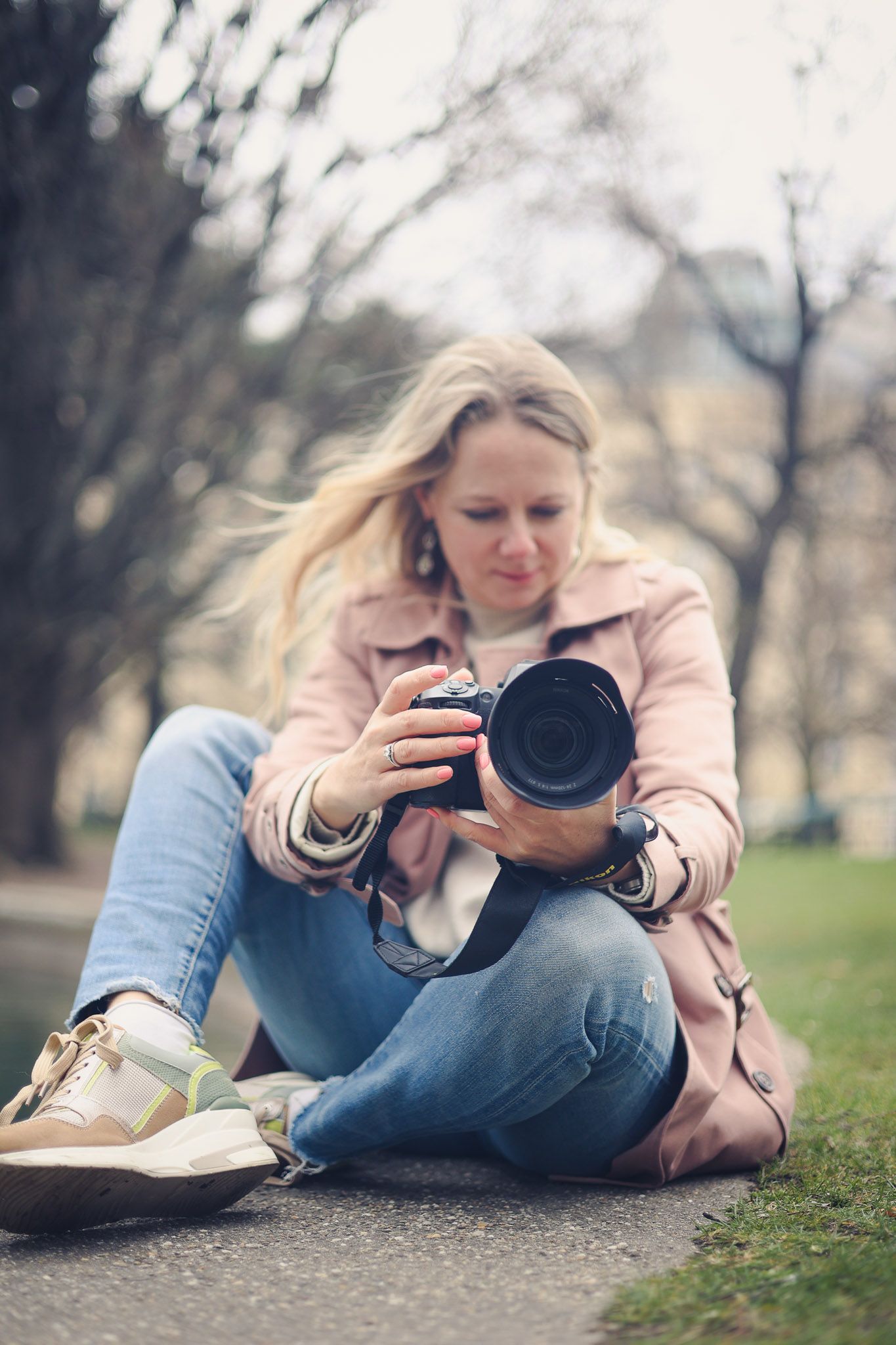 Woman in pink jacket and jeans sitting on path in park, reviewing photos on a DSLR camera.