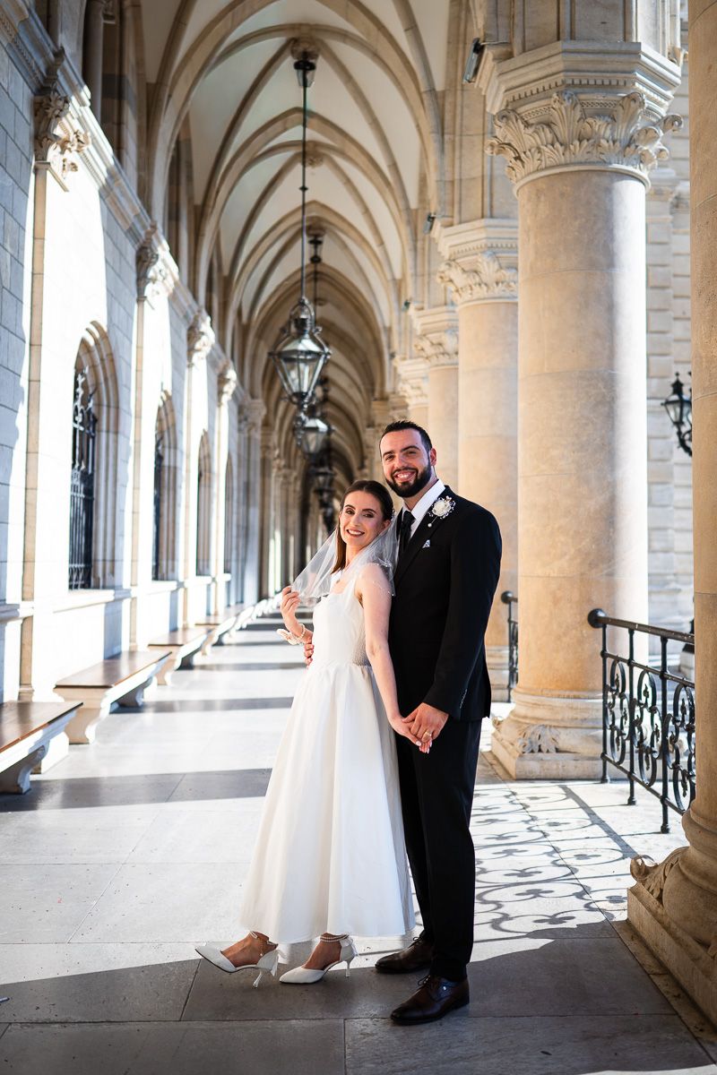 Bride and groom pose together in elegant arched hallway with marble columns on their wedding day.