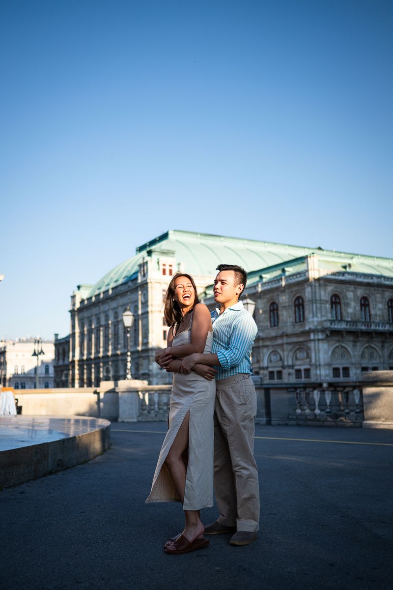 Happy couple embracing outdoors near a grand European building with a green copper roof under a clear blue sky.
