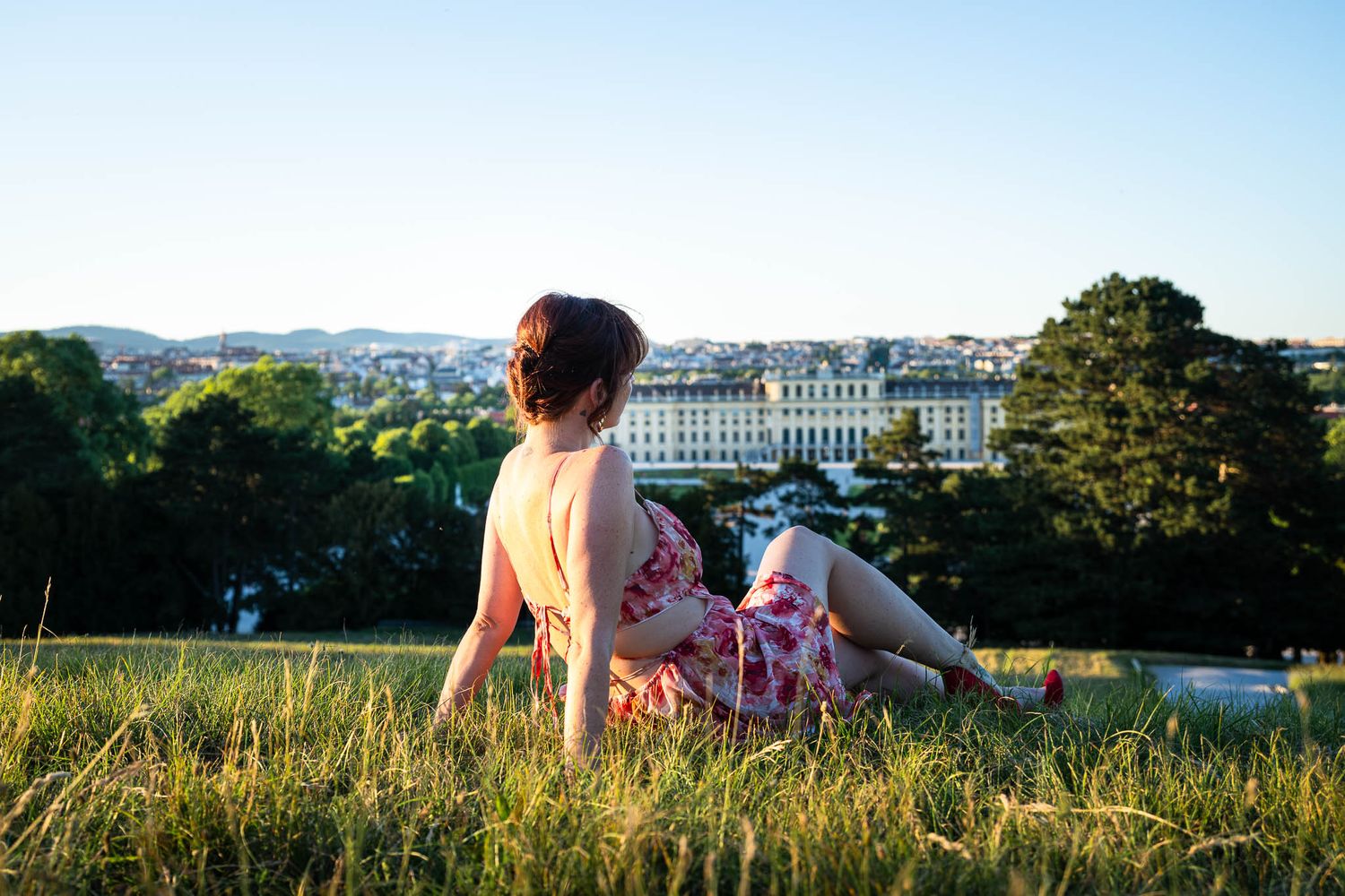 Woman in floral dress relaxing on grass hill overlooking Schönbrunn Palace in Vienna on a sunny day.