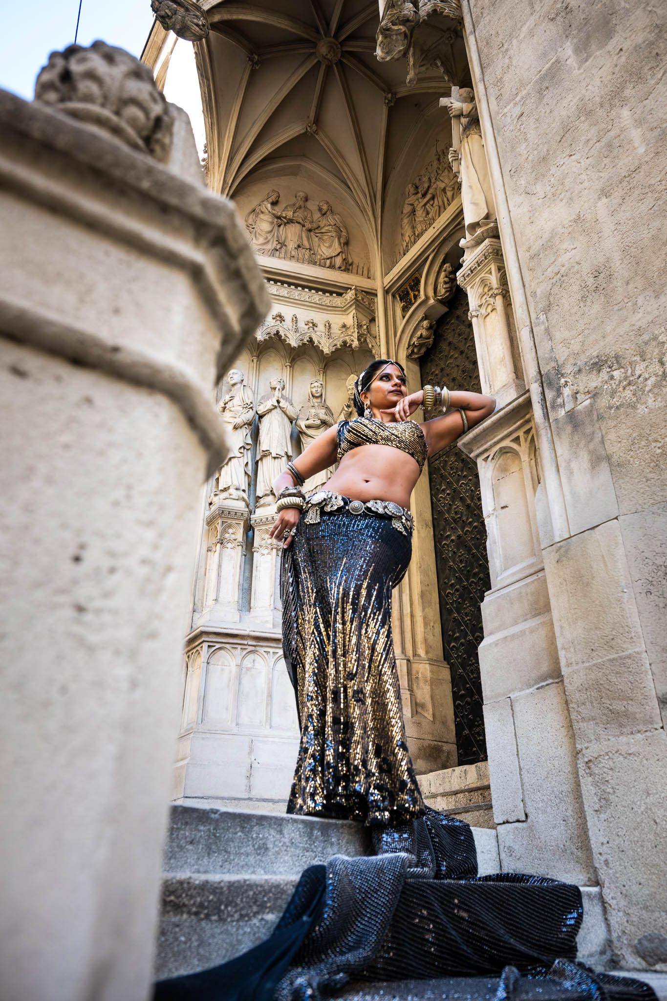 Woman in ornate gold Indian attire poses gracefully on stone steps of an elaborate Gothic cathedral.