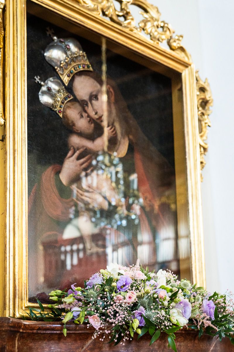 A gilded framed painting of the Virgin Mary and Christ child with silver crowns, adorned with a bouquet of purple flowers.