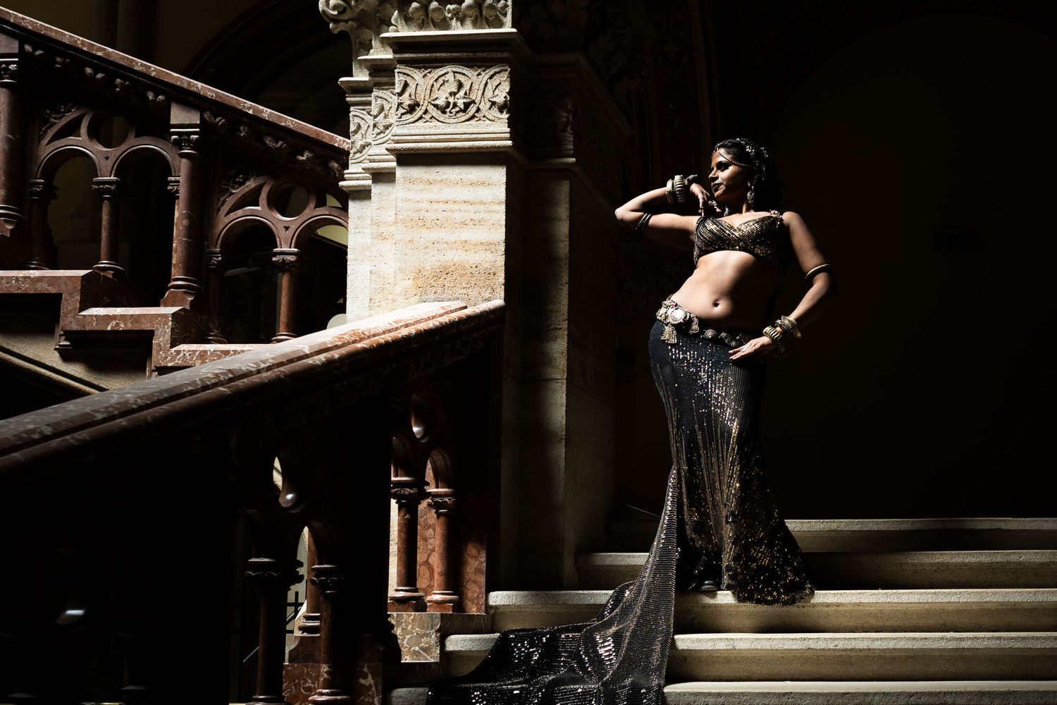 Elegant woman in ornate beaded sari poses dramatically on grand stone staircase with classical columns.