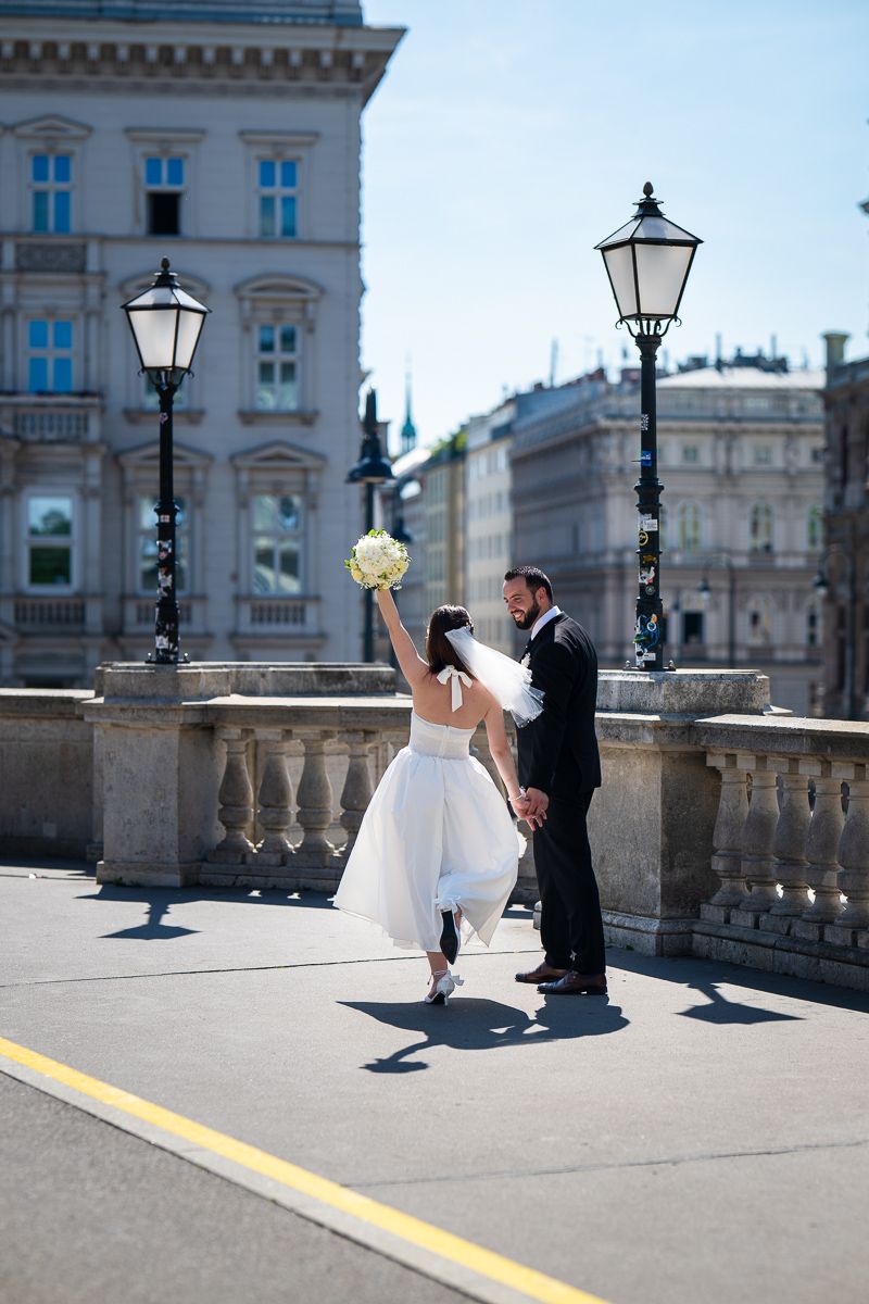 Bride and groom celebrate on a historic European bridge, bride raising bouquet, with ornate lampposts and classical buildings.