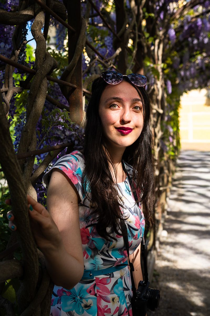 Young woman with red lips and sunglasses poses among purple wisteria flowers wearing a floral dress with a camera.