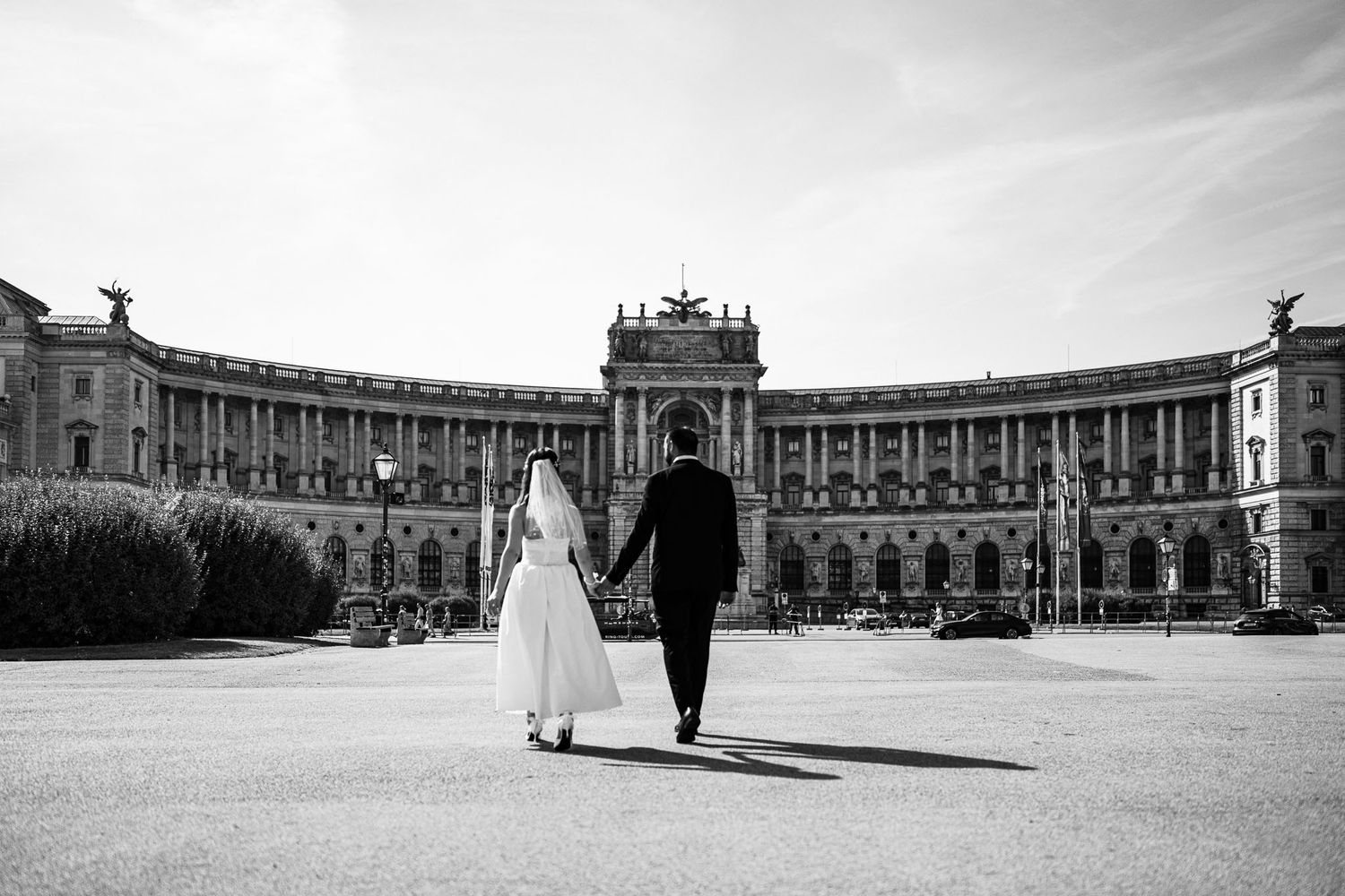Black and white photo of a bride and groom walking hand in hand toward a grand European palace.