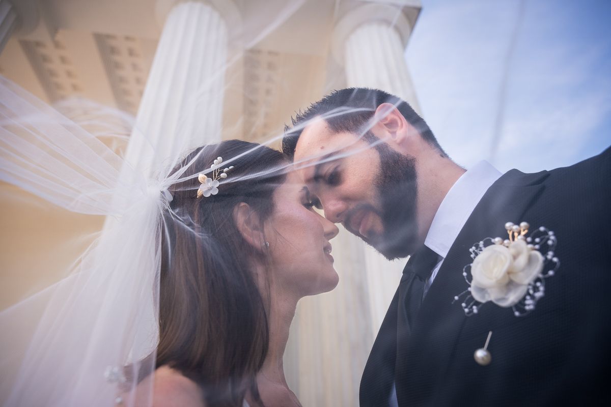 Bride and groom share an intimate moment under a flowing veil at a classic white columned wedding venue.