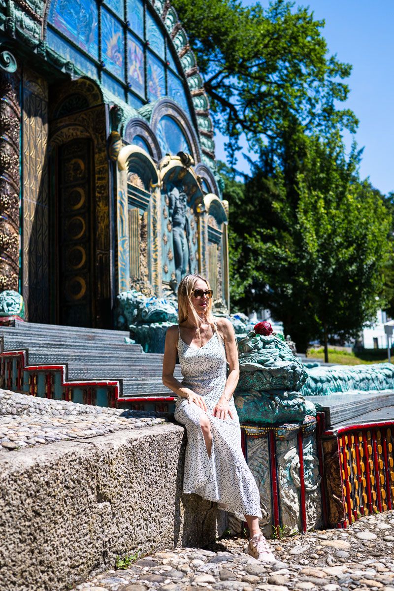 A woman in a white sundress and sunglasses sits on stone steps before an ornate Art Nouveau building facade.