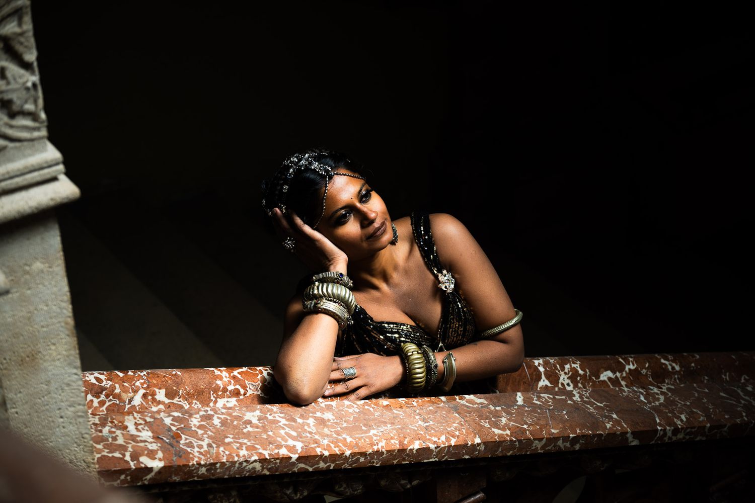 A woman wearing bangles rests her chin on her hand, leaning on a marble ledge in dramatic lighting.