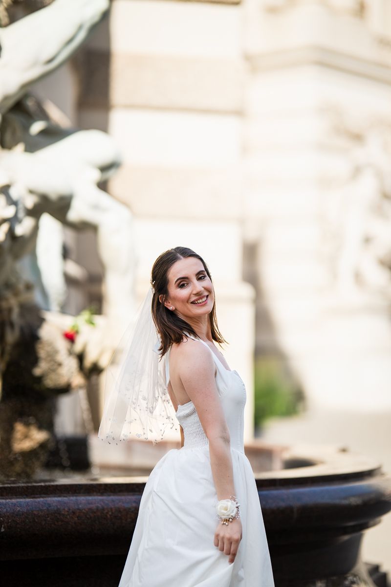 Smiling bride in white gown and veil poses gracefully by an ornate stone fountain outdoors.