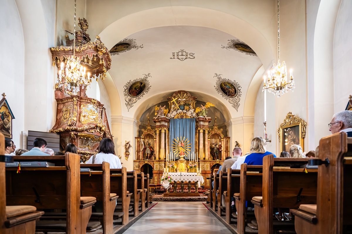 Interior of an ornate Baroque church with golden altar, wooden pews, and worshippers during a service.
