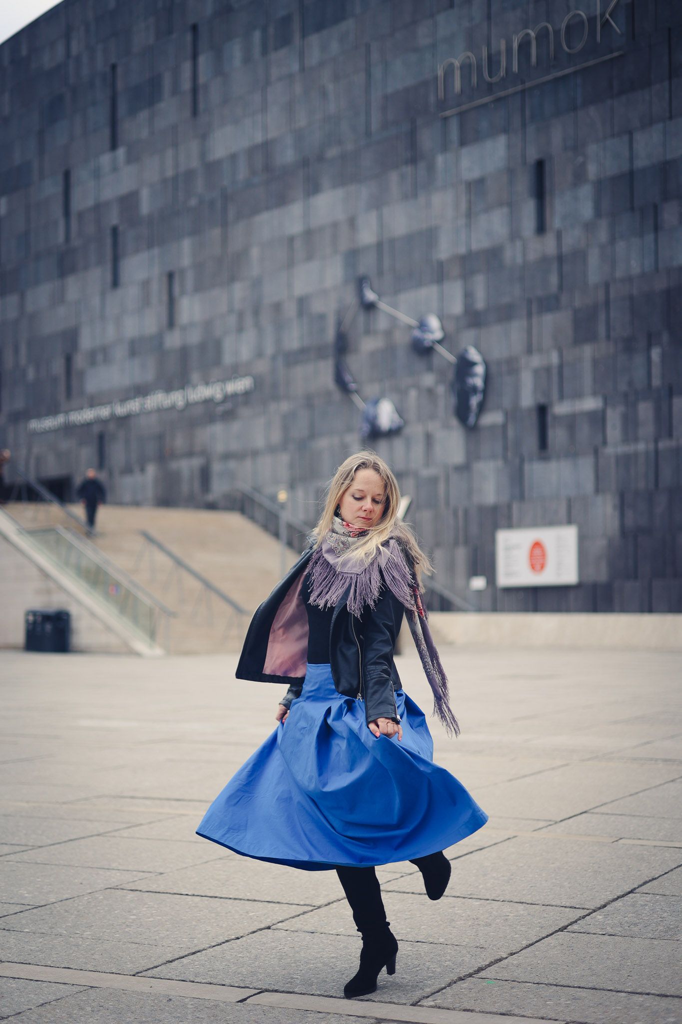 Woman in blue skirt and black jacket twirling outside the Mumok museum in Vienna, Austria.
