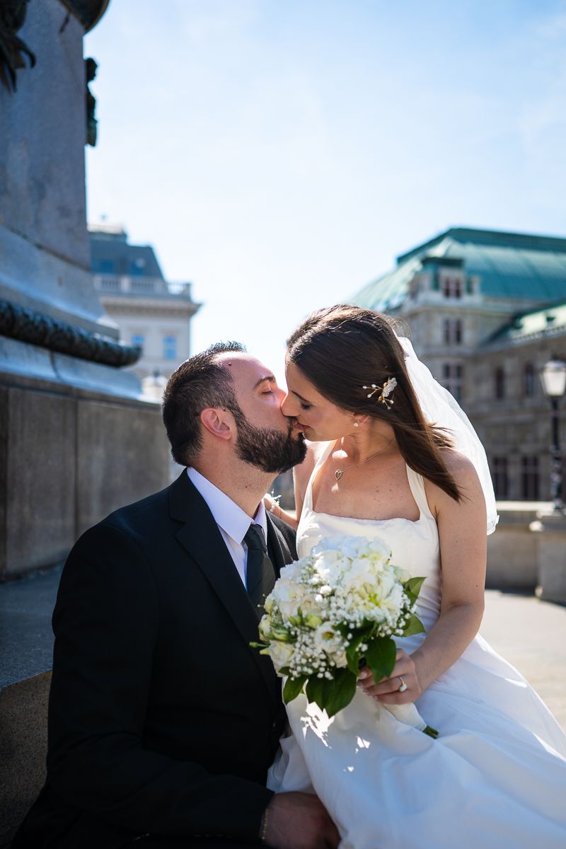 A bride and groom share a romantic kiss outdoors, she holds a white bouquet, wearing a veil, he wears a black suit.