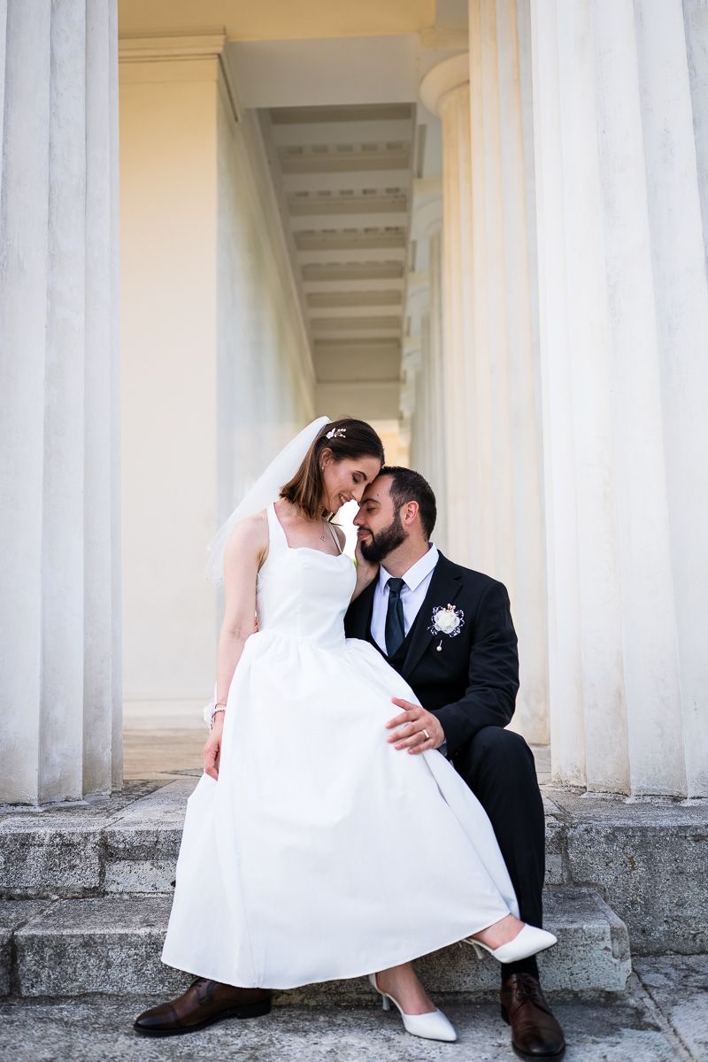 Bride in white gown and groom in black suit share a romantic moment on stone steps between tall classical columns.