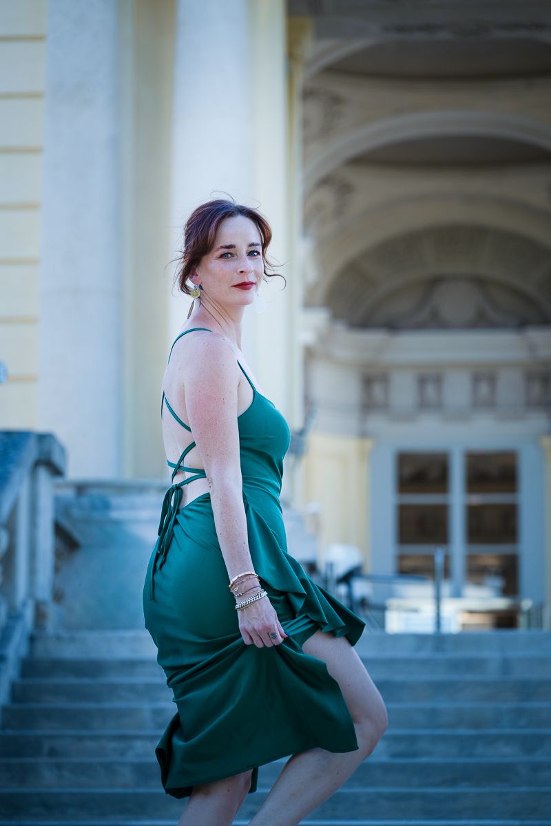 Woman in elegant green satin dress poses on grand staircase outside classical building.