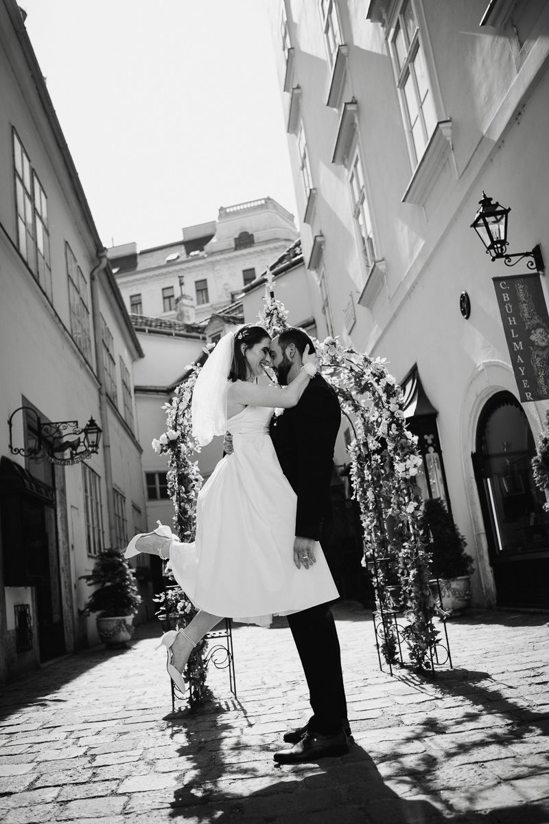 Black and white photo of a bride and groom dancing in a cobblestone alley with a floral arch backdrop.