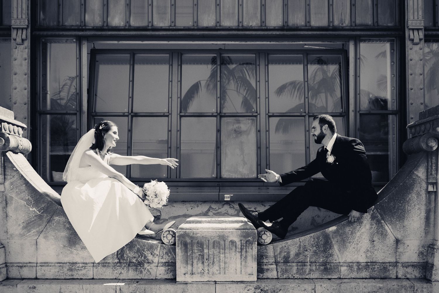 Black and white wedding photo of bride and groom sitting on stone ledge reaching toward each other.