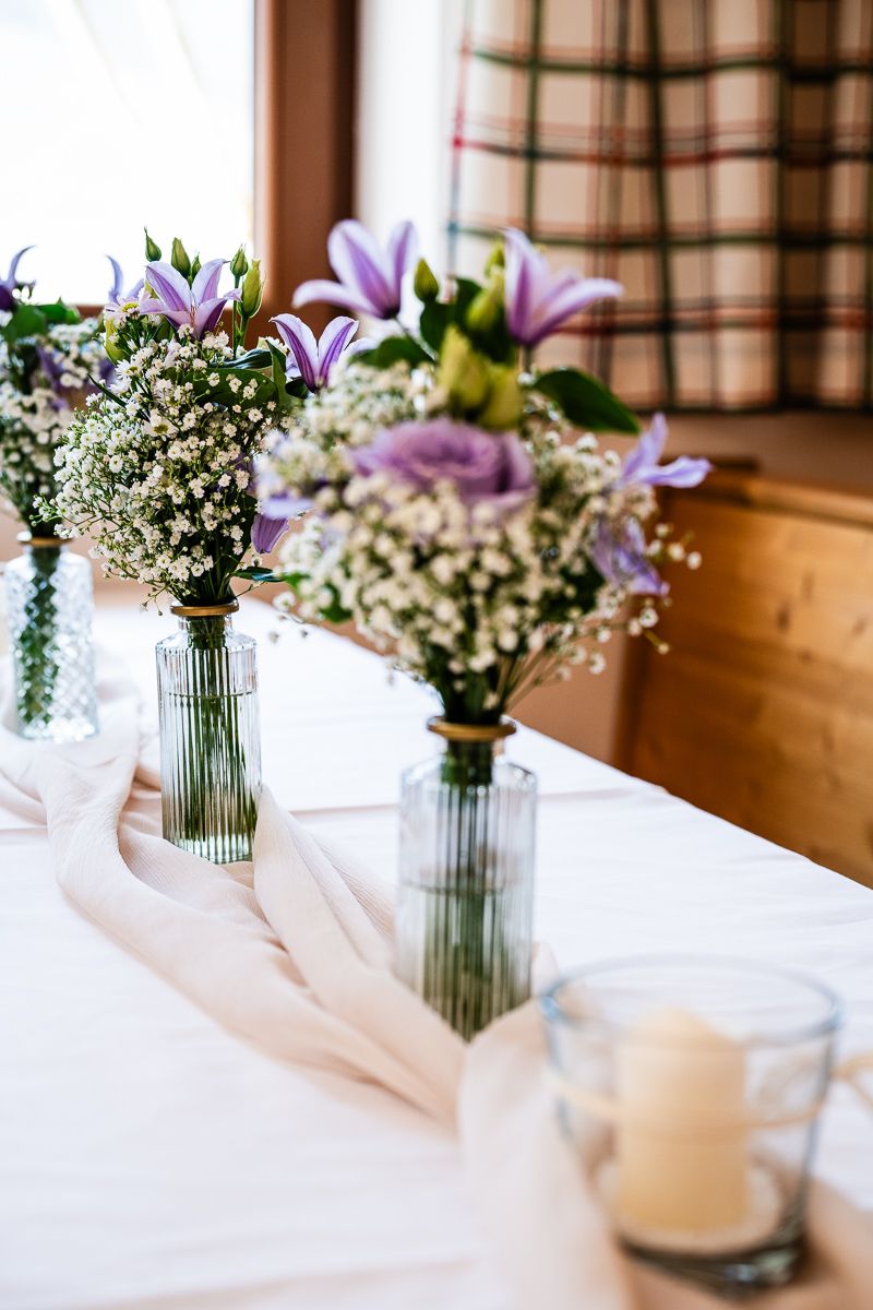 Elegant wedding table with purple floral arrangements in glass vases, white fabric runner, and candles near a window.