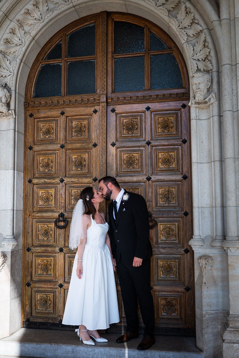 Bride and groom kissing in front of ornate wooden church doors, bride in white tea-length dress with veil.