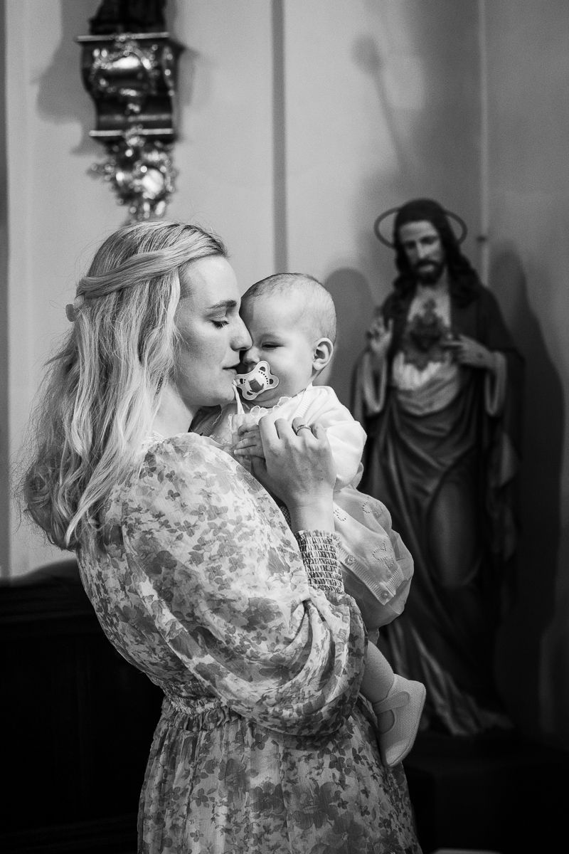 Black and white photo of a mother holding her baby in a church with a Jesus statue in the background.