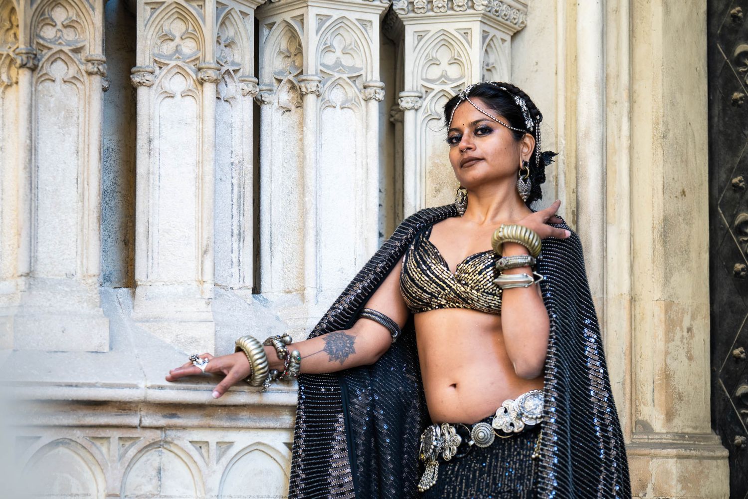 Woman in ornate black and gold Indian dance costume poses gracefully before white stone architectural columns.