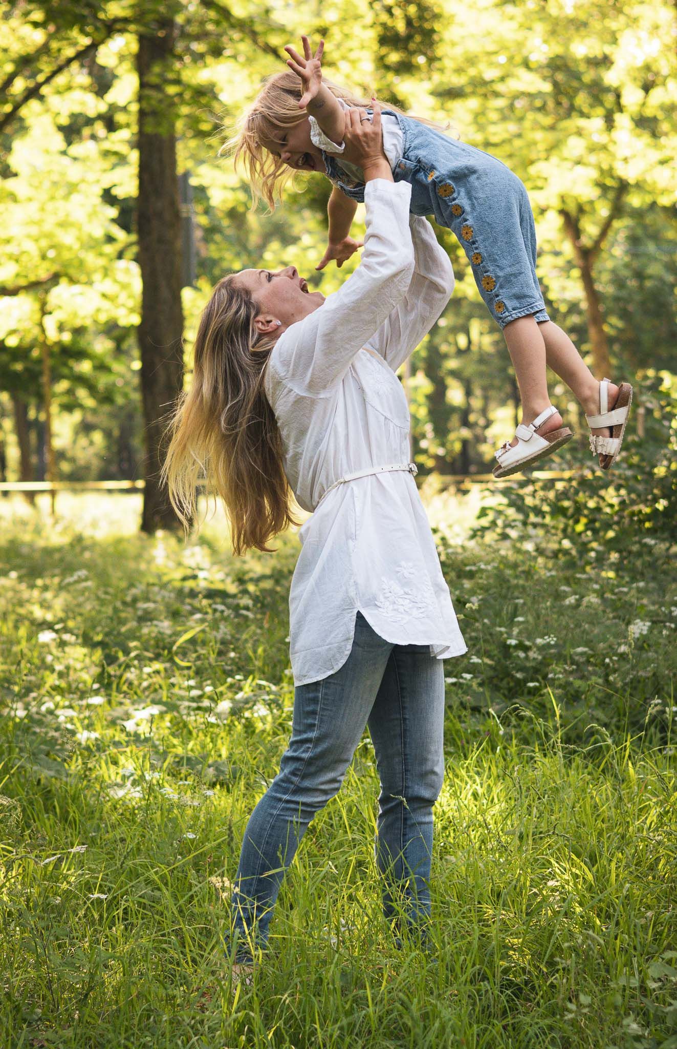 A joyful mother lifts her young child high in a sunlit forest clearing, both smiling amid lush green grass.